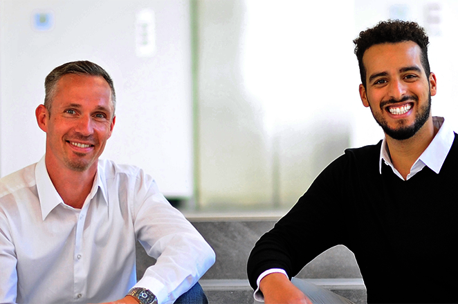 Two men are sitting next to each other on a staircase, smiling and wearing formal attire. The background is bright and modern.